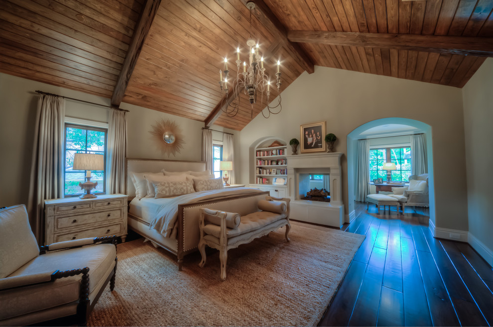 Photo of a large traditional master bedroom in Houston with white walls, dark hardwood floors, a two-sided fireplace, a wood fireplace surround and brown floor.