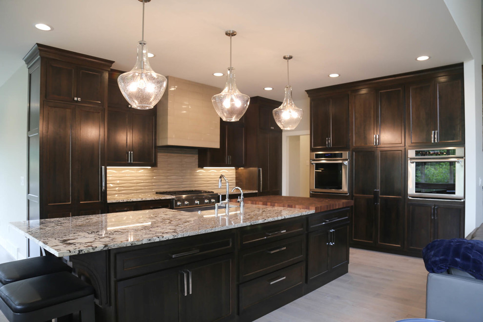 Eat-in kitchen - large transitional l-shaped light wood floor and beige floor eat-in kitchen idea with a farmhouse sink, recessed-panel cabinets, dark wood cabinets, granite countertops, gray backsplash, porcelain backsplash, stainless steel appliances, an island and multicolored countertops