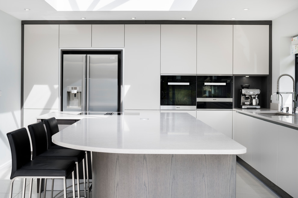 Mid-sized modern l-shaped eat-in kitchen in Sussex with a drop-in sink, flat-panel cabinets, white cabinets, quartzite benchtops, black appliances, ceramic floors, with island, white floor and grey benchtop.