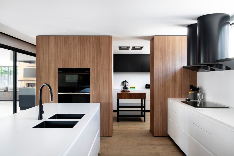 This is an example of a large contemporary l-shaped kitchen pantry in Sydney with an undermount sink, flat-panel cabinets, solid surface benchtops, white splashback, black appliances, with island, white benchtop and medium hardwood floors.