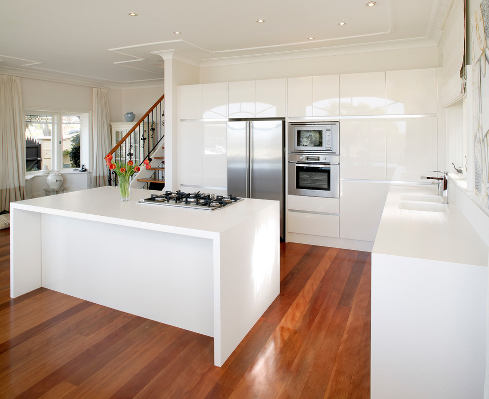 This is an example of a galley eat-in kitchen in Sydney with an integrated sink, flat-panel cabinets, white cabinets, solid surface benchtops, stainless steel appliances, medium hardwood floors and with island.