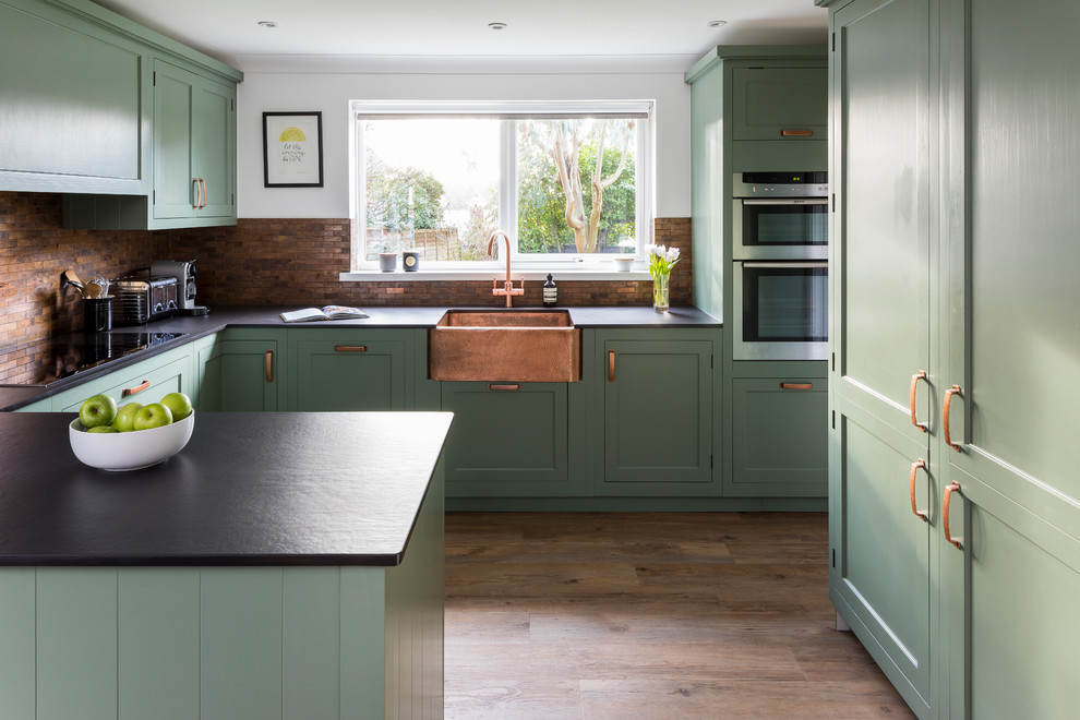 Mid-sized traditional u-shaped eat-in kitchen in Kent with a farmhouse sink, shaker cabinets, green cabinets, quartz benchtops, metal splashback, linoleum floors, a peninsula, brown splashback, stainless steel appliances and brown floor.