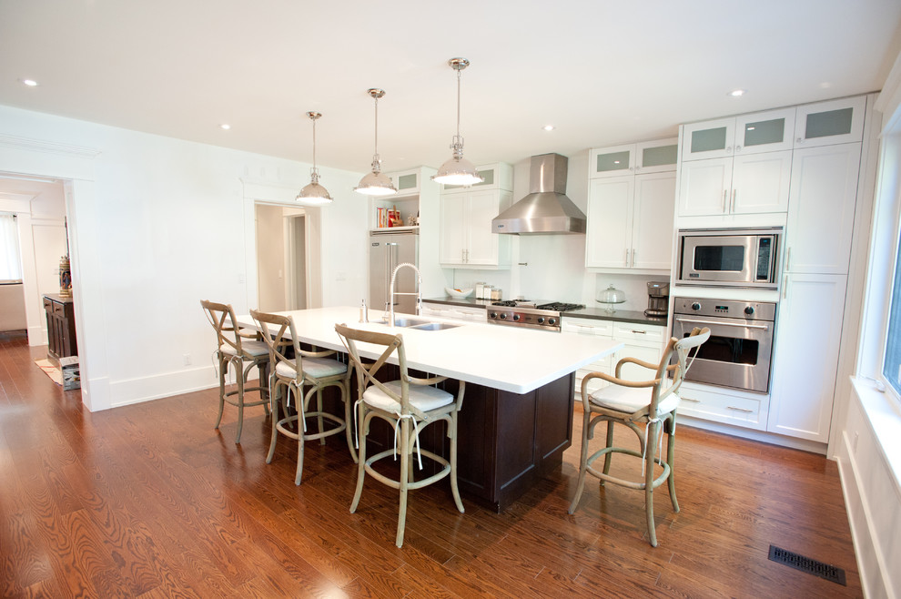 This is an example of a large beach style galley separate kitchen in Toronto with stainless steel appliances, an undermount sink, shaker cabinets, white cabinets, quartz benchtops, white splashback, subway tile splashback, medium hardwood floors, with island and brown floor.