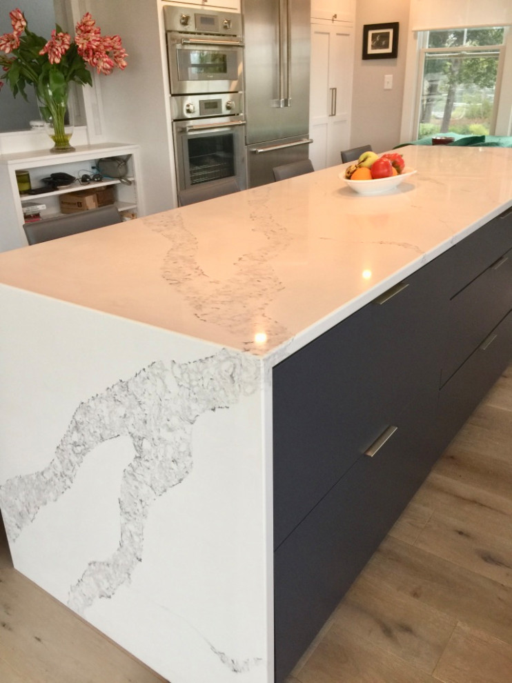 Photo of a small beach style l-shaped eat-in kitchen in Portland Maine with an undermount sink, recessed-panel cabinets, white cabinets, quartz benchtops, metallic splashback, engineered quartz splashback, stainless steel appliances, medium hardwood floors, with island, brown floor, white benchtop and timber.