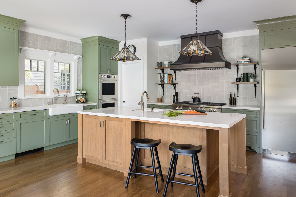 Large classic l-shaped kitchen/diner in Los Angeles with a belfast sink, shaker cabinets, green cabinets, grey splashback, metro tiled splashback, stainless steel appliances, medium hardwood flooring, an island, brown floors and multicoloured worktops.