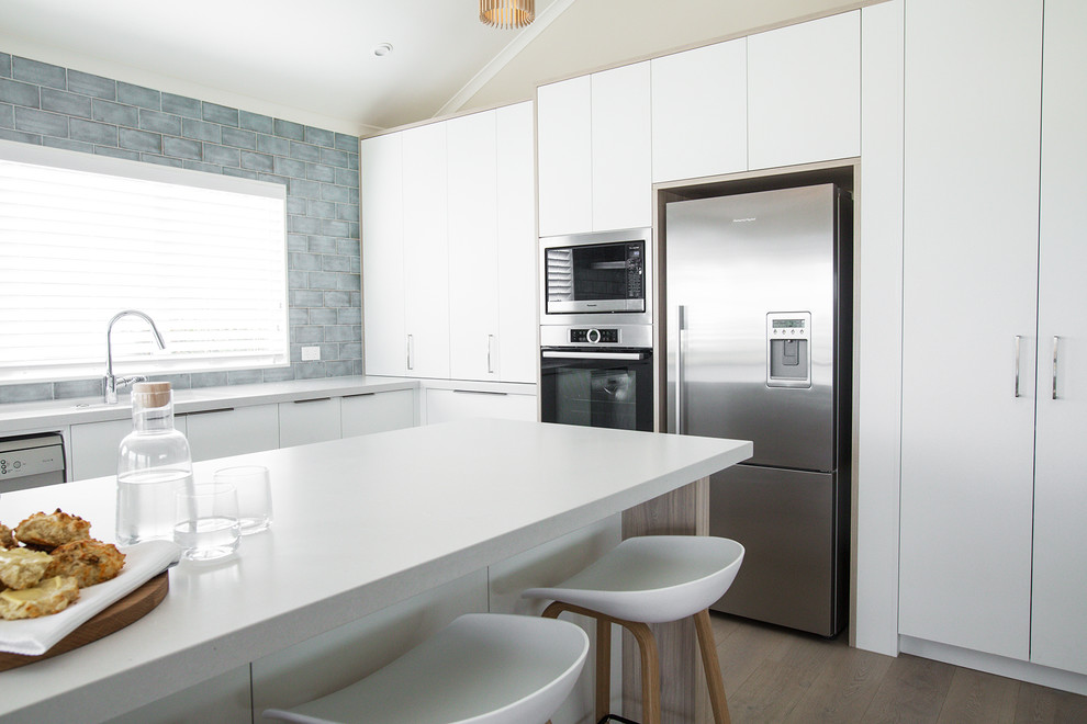 Large transitional eat-in kitchen in Auckland with white cabinets, blue splashback, ceramic splashback, stainless steel appliances, laminate floors, with island, brown floor and white benchtop.