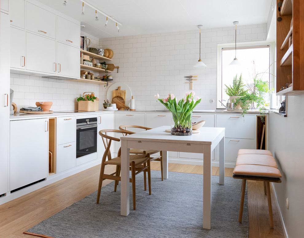 Photo of a mid-sized scandinavian l-shaped eat-in kitchen in Malmo with flat-panel cabinets, white cabinets, quartz benchtops, white splashback, subway tile splashback, black appliances, light hardwood floors and brown floor.