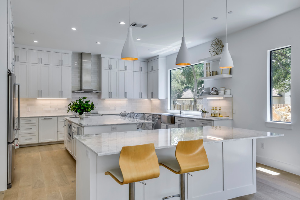 Photo of a large contemporary u-shaped open plan kitchen in Austin with shaker cabinets, white cabinets, white splashback, light hardwood floors, multiple islands, beige floor, a farmhouse sink, quartzite benchtops, stone tile splashback, stainless steel appliances and white benchtop.