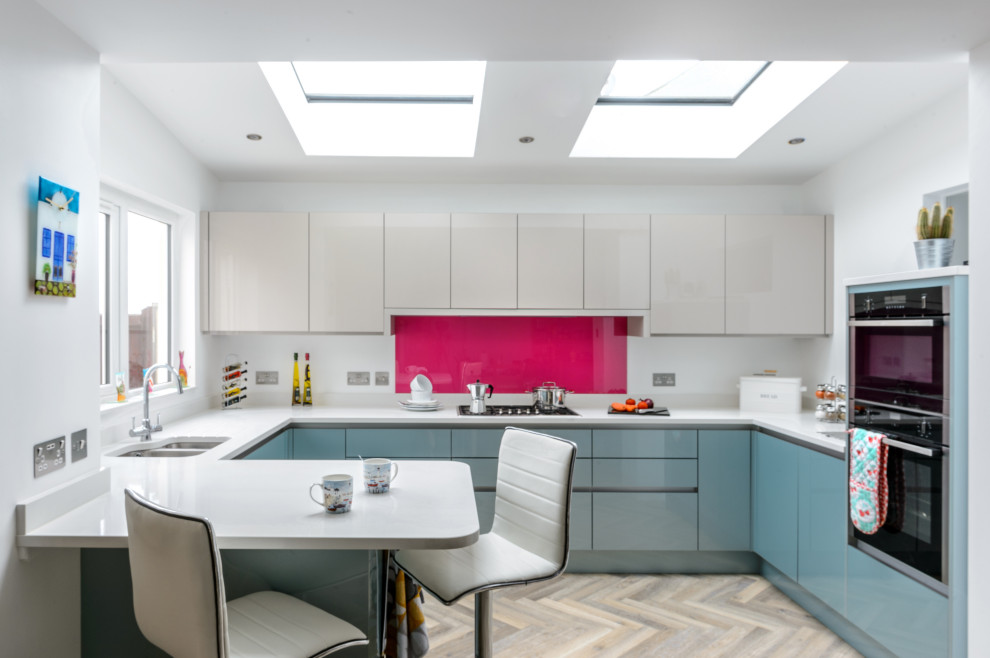Mid-sized modern l-shaped open plan kitchen in Cornwall with a drop-in sink, glass-front cabinets, beige cabinets, pink splashback, glass sheet splashback, black appliances, light hardwood floors, with island and beige floor.