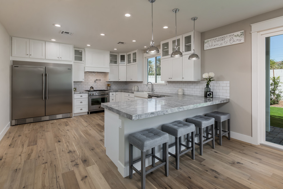 Transitional l-shaped kitchen in Phoenix with a farmhouse sink, shaker cabinets, white cabinets, white splashback, subway tile splashback, stainless steel appliances, a peninsula, brown floor and grey benchtop.