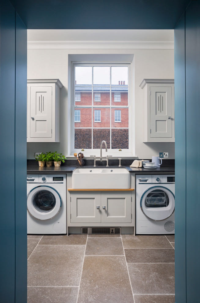 This is an example of a mid-sized transitional laundry room in Hampshire with grey cabinets, granite benchtops, black splashback, limestone floors, shaker cabinets and a farmhouse sink.