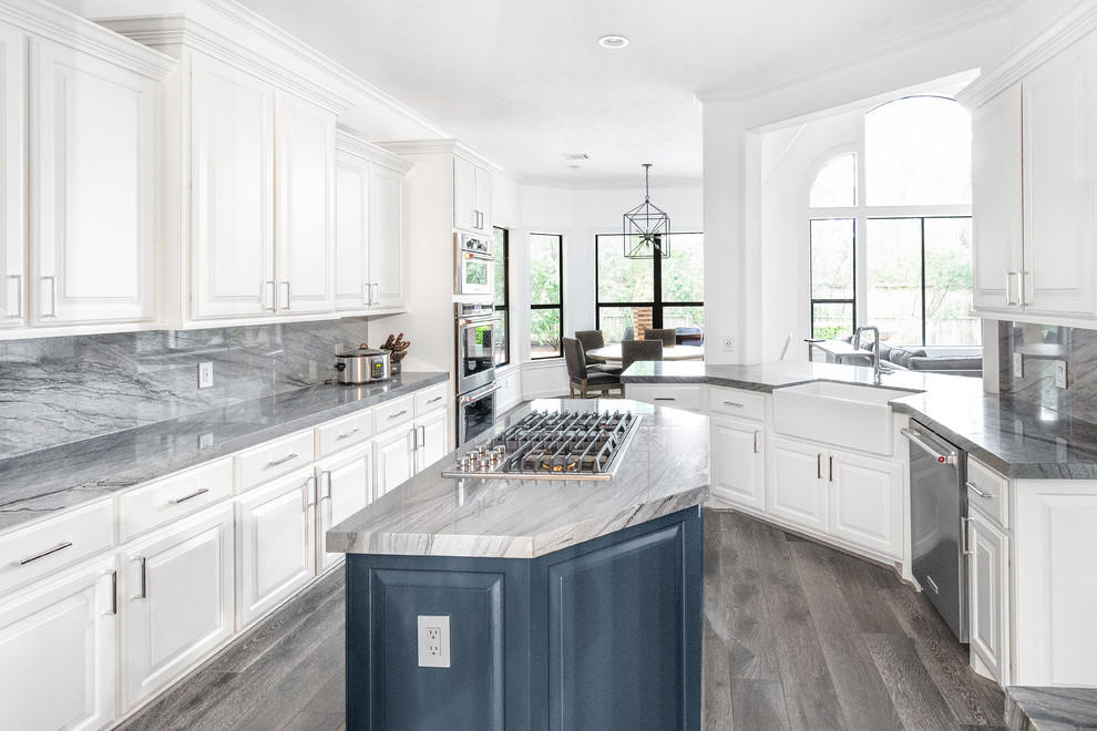 This is an example of a mid-sized transitional galley open plan kitchen in Houston with a farmhouse sink, raised-panel cabinets, blue cabinets, quartzite benchtops, grey splashback, stone slab splashback, stainless steel appliances, medium hardwood floors, with island, grey floor and grey benchtop.