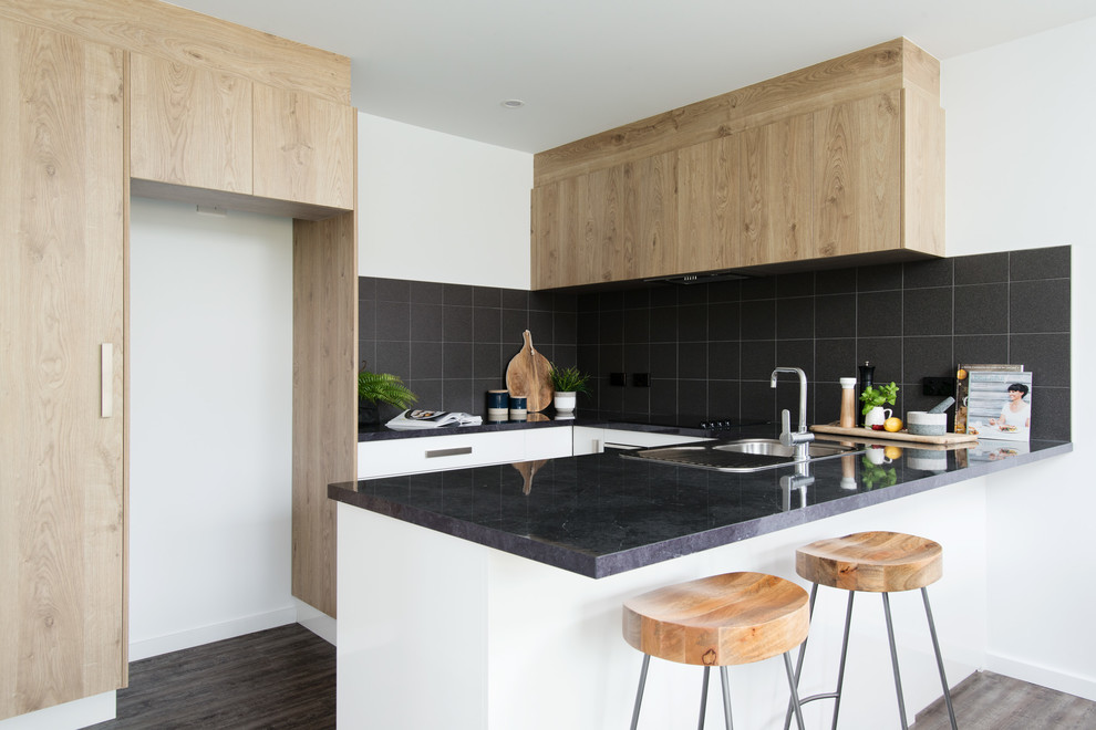 Small contemporary u-shaped open plan kitchen in Other with a drop-in sink, white cabinets, laminate benchtops, black splashback, ceramic splashback, black appliances, vinyl floors, no island, brown floor and black benchtop.