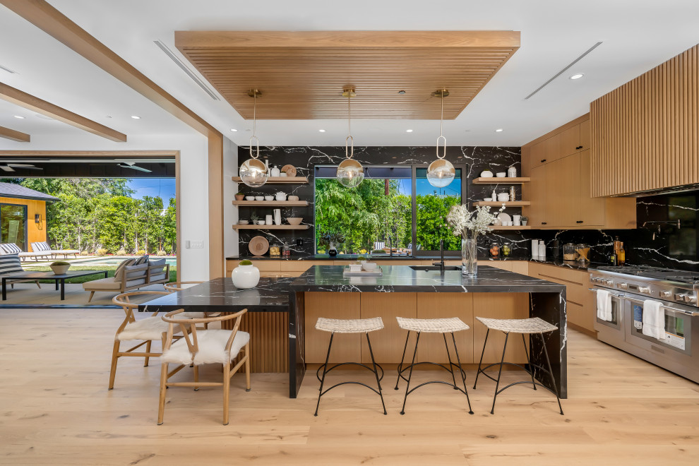 Photo of a contemporary l-shaped kitchen in Los Angeles with an undermount sink, flat-panel cabinets, medium wood cabinets, black splashback, stone slab splashback, stainless steel appliances, light hardwood floors, with island, beige floor and black benchtop.
