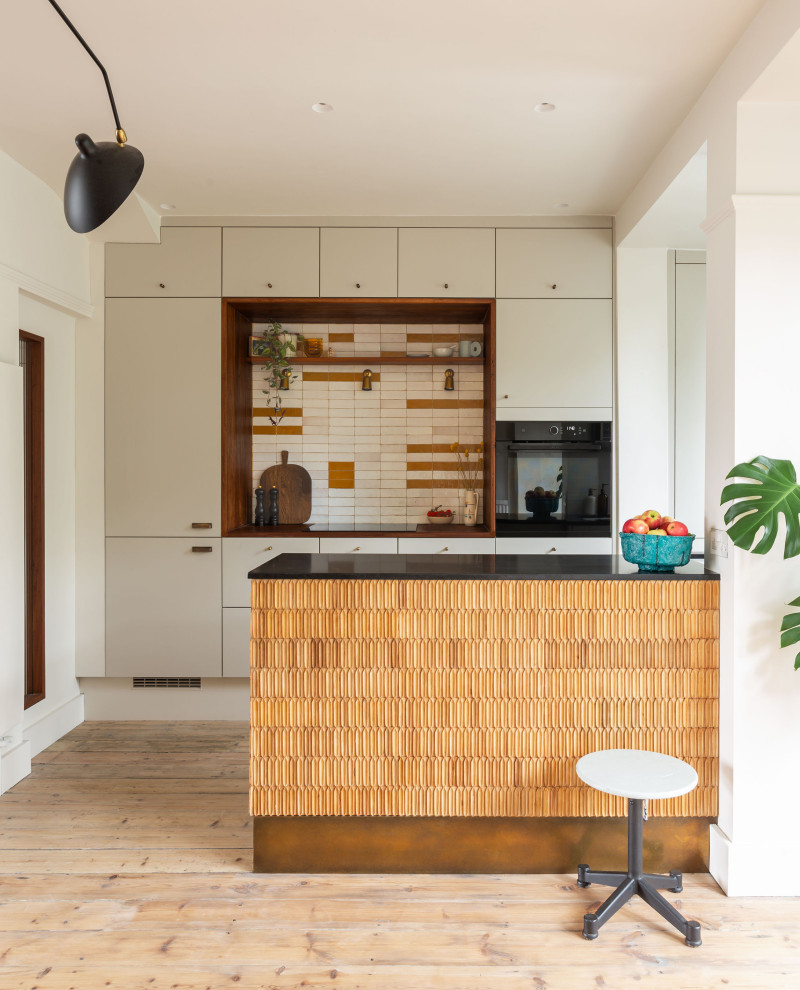 Photo of an eclectic galley kitchen in London with flat-panel cabinets, grey cabinets, multi-coloured splashback, black appliances, medium hardwood flooring, a breakfast bar, brown floors and black worktops.