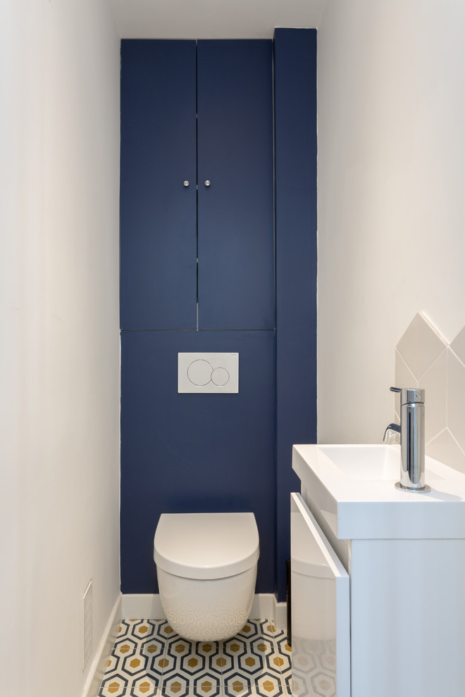 This is an example of a mid-sized modern powder room in Paris with white walls, beaded inset cabinets, blue cabinets, a wall-mount toilet, white tile, porcelain tile, cement tiles, a wall-mount sink, solid surface benchtops, multi-coloured floor and white benchtops.