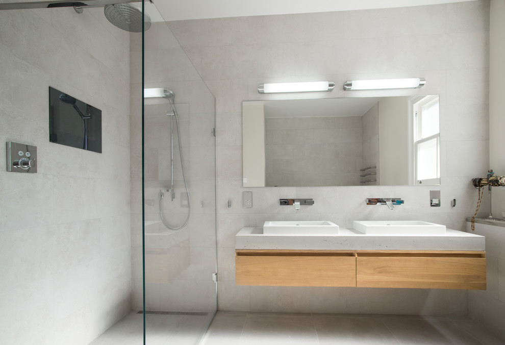 This is an example of a mid-sized contemporary master wet room bathroom in London with flat-panel cabinets, light wood cabinets, grey walls, a vessel sink, an open shower, an alcove tub, a wall-mount toilet, beige tile, travertine, ceramic floors, concrete benchtops and beige floor.