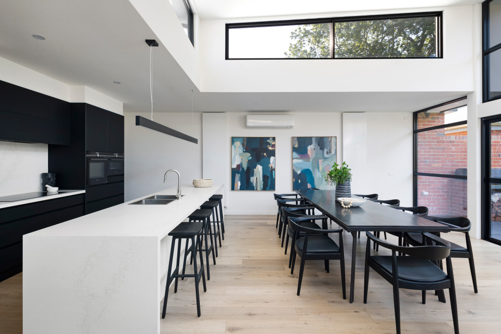 Inspiration for a mid-sized modern galley open plan kitchen in Melbourne with an undermount sink, flat-panel cabinets, black cabinets, quartz benchtops, white splashback, glass sheet splashback, black appliances, light hardwood floors, with island, brown floor and white benchtop.
