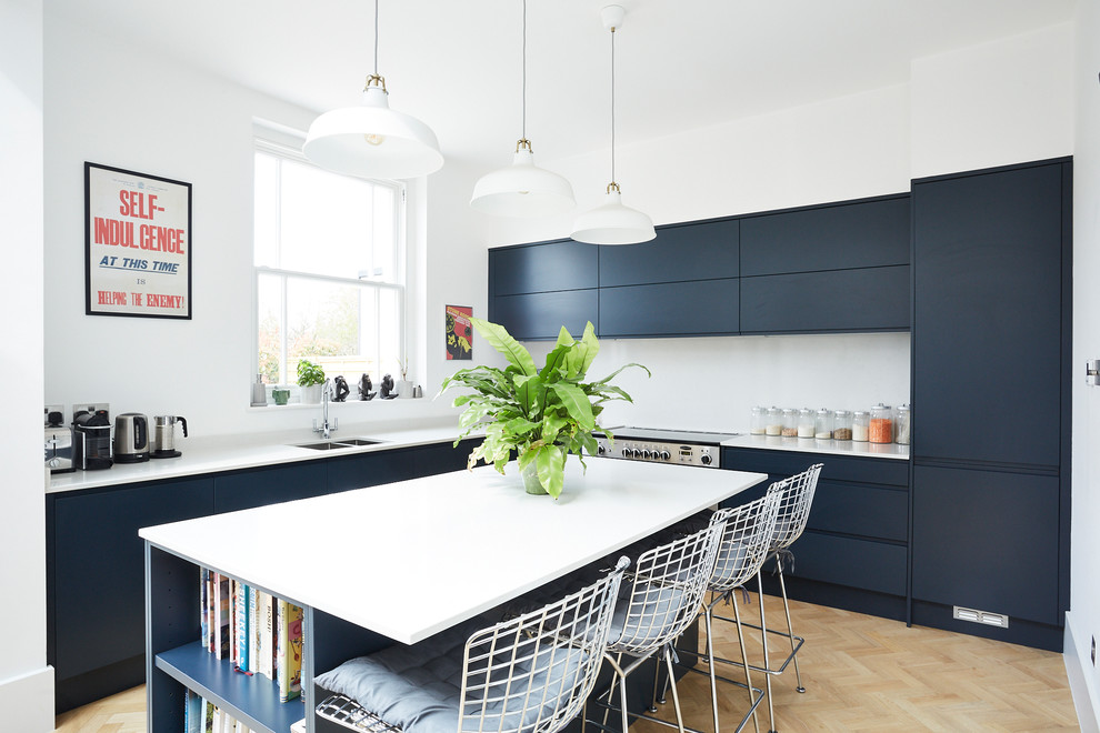 Photo of a contemporary l-shaped kitchen in London with an undermount sink, flat-panel cabinets, blue cabinets, stainless steel appliances, light hardwood floors, with island, beige floor and white benchtop.