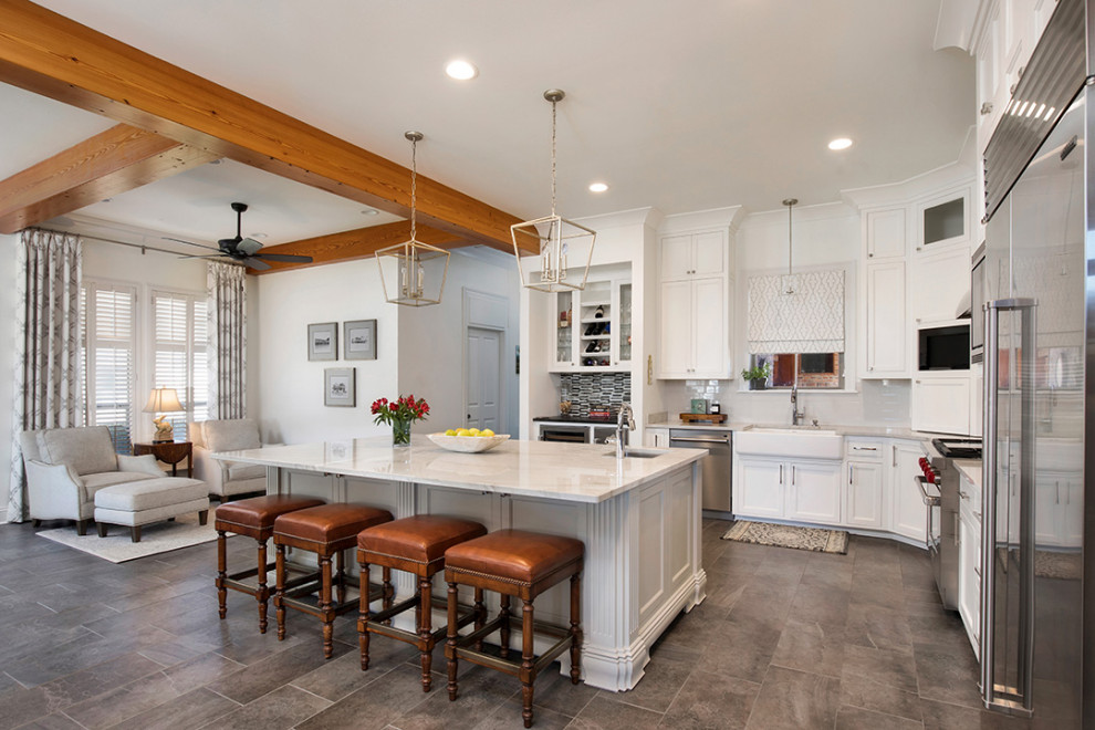 Inspiration for a mid-sized transitional l-shaped eat-in kitchen in New Orleans with a farmhouse sink, recessed-panel cabinets, white cabinets, quartzite benchtops, grey splashback, ceramic splashback, stainless steel appliances, porcelain floors, with island, grey floor, white benchtop and exposed beam.