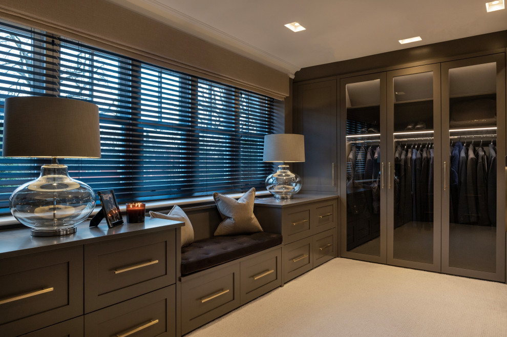 Design ideas for a large transitional men's walk-in wardrobe in Surrey with glass-front cabinets, brown cabinets and carpet.