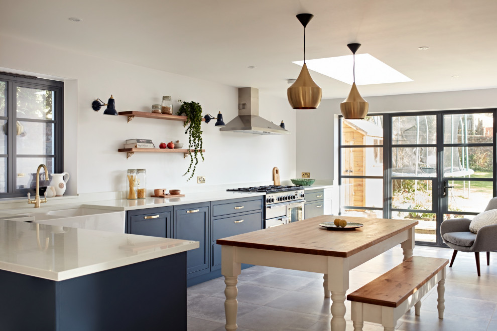 This is an example of a large classic kitchen/diner in Sussex with a belfast sink, shaker cabinets, blue cabinets, marble worktops, white splashback, marble splashback, stainless steel appliances, porcelain flooring, a breakfast bar, grey floors, white worktops and feature lighting.