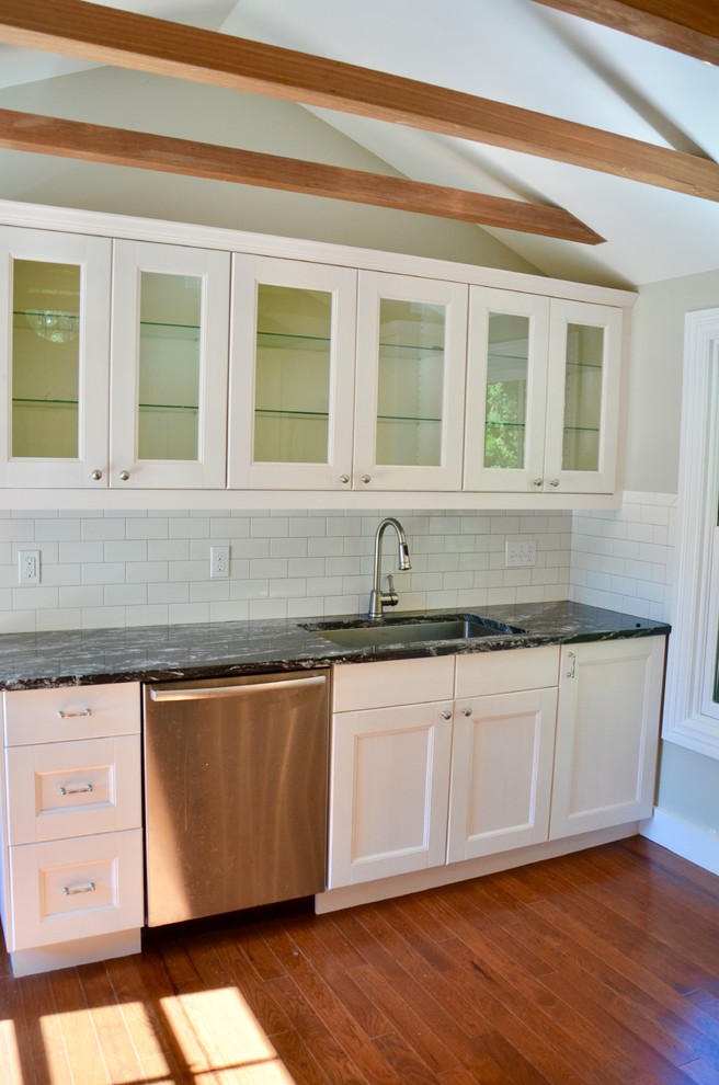 Small transitional medium tone wood floor enclosed kitchen photo with an undermount sink, glass-front cabinets, granite countertops, white backsplash, ceramic backsplash and stainless steel appliances