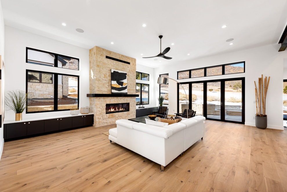 На фото: большая открытая гостиная комната в стиле модернизм с белыми стенами, светлым паркетным полом, стандартным камином, фасадом камина из камня, телевизором на стене, коричневым полом и балками на потолке с