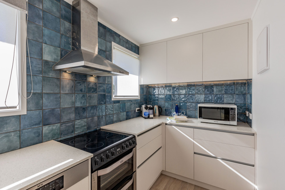This is an example of a large contemporary u-shaped kitchen/diner in Auckland with a double-bowl sink, flat-panel cabinets, white cabinets, composite countertops, blue splashback, ceramic splashback, stainless steel appliances, laminate floors, a breakfast bar, brown floors and white worktops.