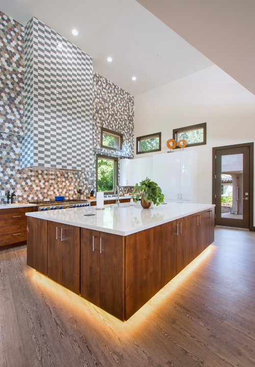 Modern kitchen with a large wood island and quartz top, LED toe-kick accent lighting, recessed ceiling lights, and a patterned tile wall with an oversized range hood