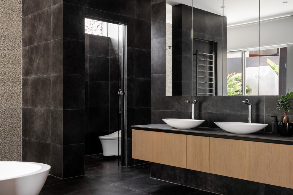 Design ideas for a contemporary master bathroom in Other with flat-panel cabinets, medium wood cabinets, a freestanding tub, a wall-mount toilet, black tile, white walls, a vessel sink, black floor, grey benchtops, an enclosed toilet, a double vanity and a floating vanity.
