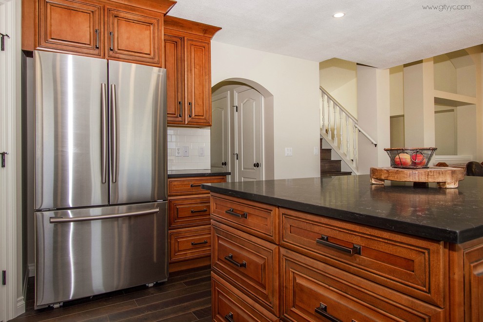 Eat-in kitchen - mid-sized rustic l-shaped laminate floor and black floor eat-in kitchen idea in Calgary with a drop-in sink, raised-panel cabinets, medium tone wood cabinets, quartzite countertops, white backsplash, ceramic backsplash, stainless steel appliances and an island