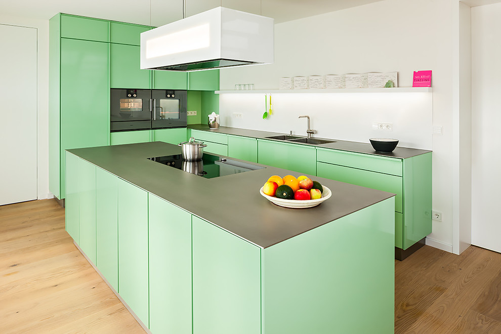 Photo of a large contemporary l-shaped separate kitchen in Berlin with a double-bowl sink, flat-panel cabinets, green cabinets, stainless steel benchtops, white splashback, panelled appliances, medium hardwood floors and with island.