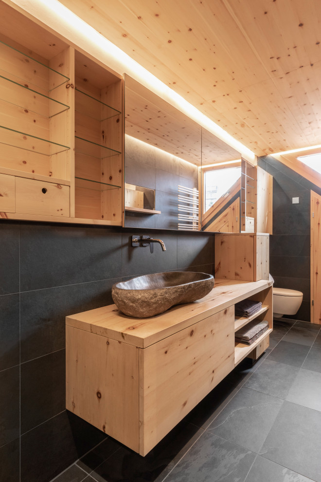 This is an example of a small contemporary bathroom in Other with flat-panel cabinets, light wood cabinets, a curbless shower, a bidet, black tile, slate, black walls, slate floors, a trough sink, granite benchtops, black floor, brown benchtops, an enclosed toilet, a single vanity, a floating vanity and wood.