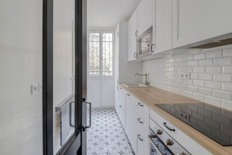Photo of a mid-sized contemporary single-wall separate kitchen in Paris with an undermount sink, shaker cabinets, white cabinets, wood benchtops, white splashback, ceramic splashback, stainless steel appliances, ceramic floors, no island, multi-coloured floor and brown benchtop.