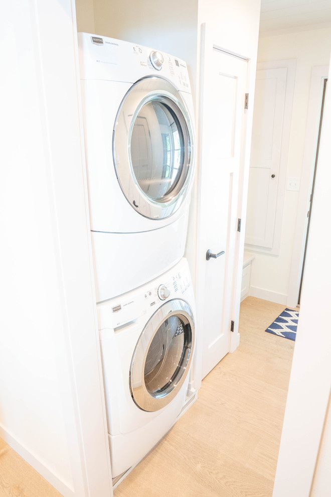 This is an example of a mid-sized beach style single-wall dedicated laundry room in Other with white walls, light hardwood floors, beige floor and a stacked washer and dryer.
