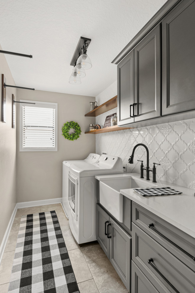 Photo of a transitional laundry room in Orlando with a farmhouse sink, recessed-panel cabinets, grey cabinets, glass benchtops, white splashback, ceramic splashback, beige walls, a side-by-side washer and dryer and white benchtop.