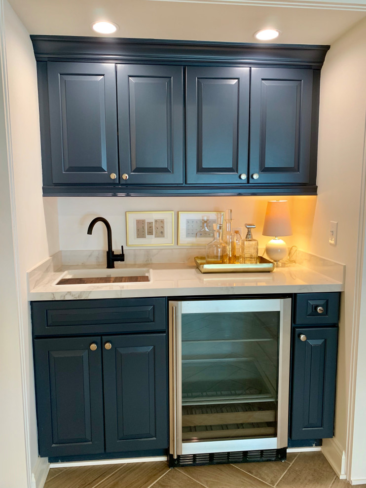 This is an example of a mid-sized transitional galley wet bar in Milwaukee with an undermount sink, raised-panel cabinets, blue cabinets, marble benchtops, white splashback, porcelain floors, brown floor and white benchtop.