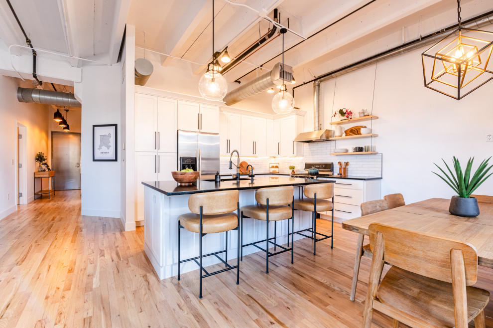 Inspiration for a large urban l-shaped kitchen/diner in Denver with an integrated sink, shaker cabinets, white cabinets, soapstone worktops, white splashback, ceramic splashback, stainless steel appliances, light hardwood flooring, an island, black worktops and exposed beams.