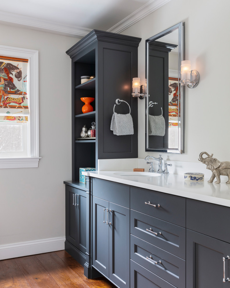 Photo of a transitional master bathroom in New York with grey cabinets, medium hardwood floors, an undermount sink, engineered quartz benchtops, white benchtops, recessed-panel cabinets, grey walls and brown floor.