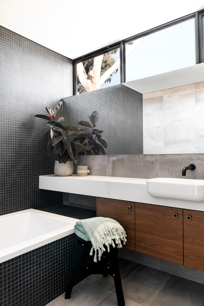 This is an example of a mid-sized contemporary kids bathroom in Perth with medium wood cabinets, a drop-in tub, black tile, porcelain tile, black walls, porcelain floors, engineered quartz benchtops, grey floor, white benchtops and a drop-in sink.