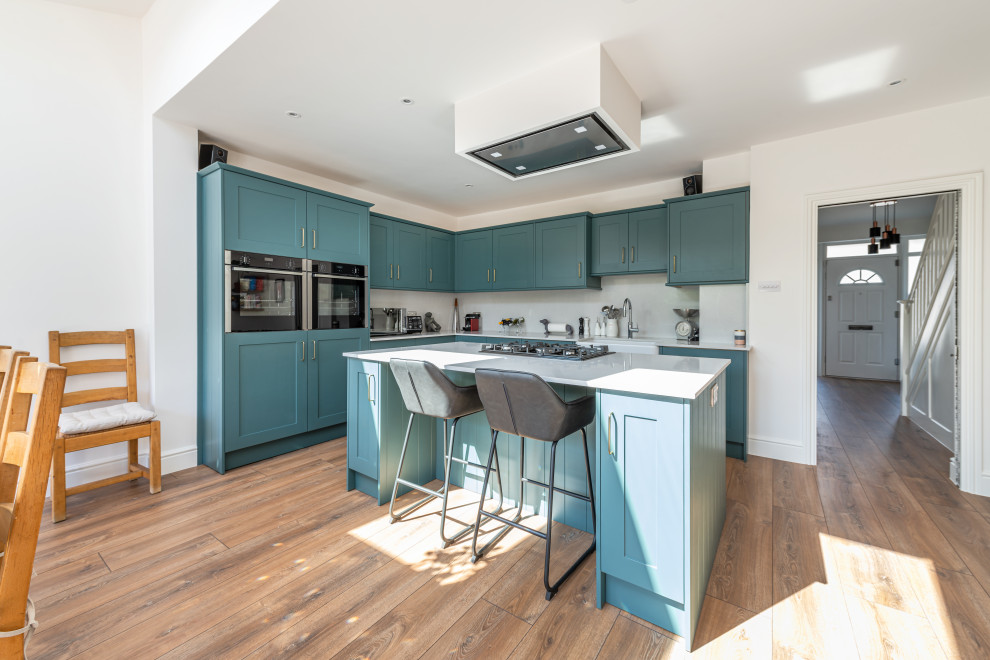 Large modern l-shaped open plan kitchen in London with a drop-in sink, shaker cabinets, turquoise cabinets, granite benchtops, white splashback, glass sheet splashback, stainless steel appliances, medium hardwood floors, with island, brown floor and white benchtop.