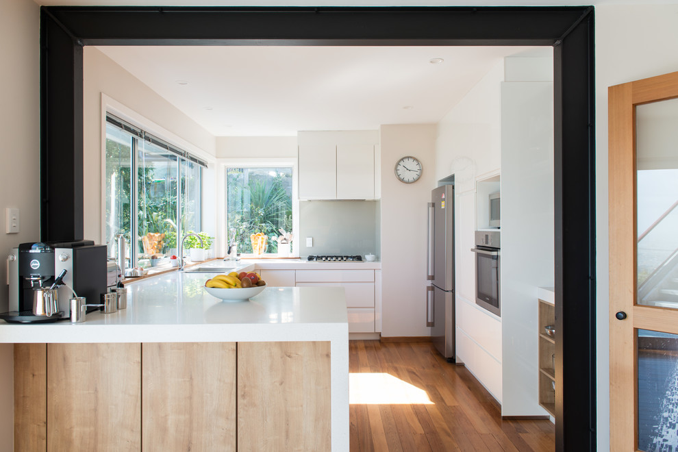 Photo of a mid-sized scandinavian u-shaped kitchen pantry in Christchurch with an undermount sink, flat-panel cabinets, white cabinets, solid surface benchtops, glass sheet splashback, white appliances, medium hardwood floors, a peninsula and brown floor.