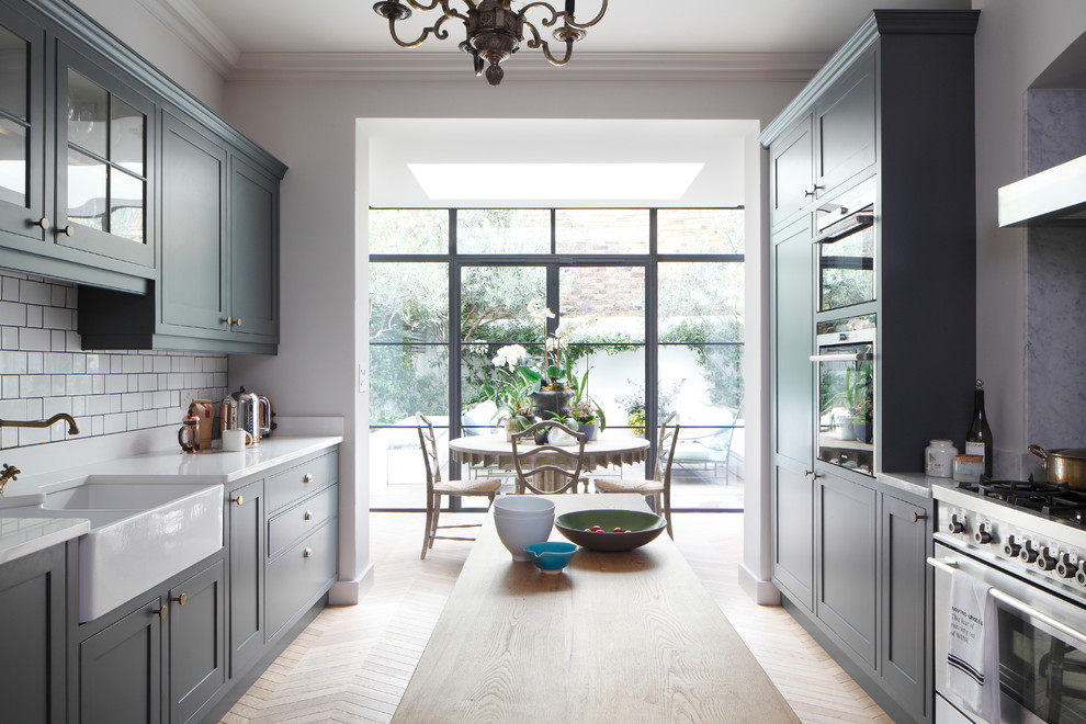 Design ideas for a large traditional galley open plan kitchen in London with a farmhouse sink, shaker cabinets, grey cabinets, quartzite benchtops, white splashback, ceramic splashback, stainless steel appliances, light hardwood floors, a peninsula, brown floor and white benchtop.