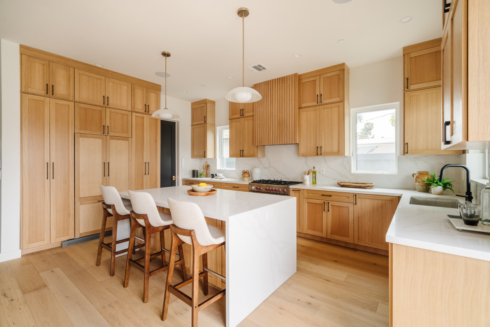 Example of a transitional u-shaped light wood floor and beige floor kitchen design in Los Angeles with shaker cabinets, light wood cabinets, an island, white countertops, an undermount sink, white backsplash, stone slab backsplash and stainless steel appliances