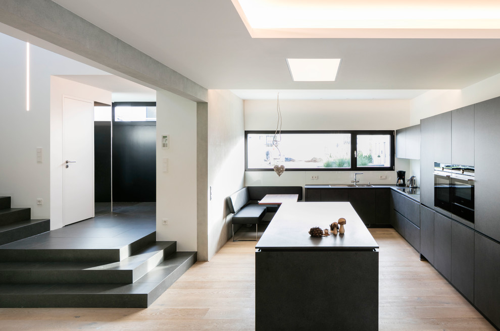 Expansive contemporary u-shaped open plan kitchen in Cologne with flat-panel cabinets, black appliances, light hardwood floors and with island.