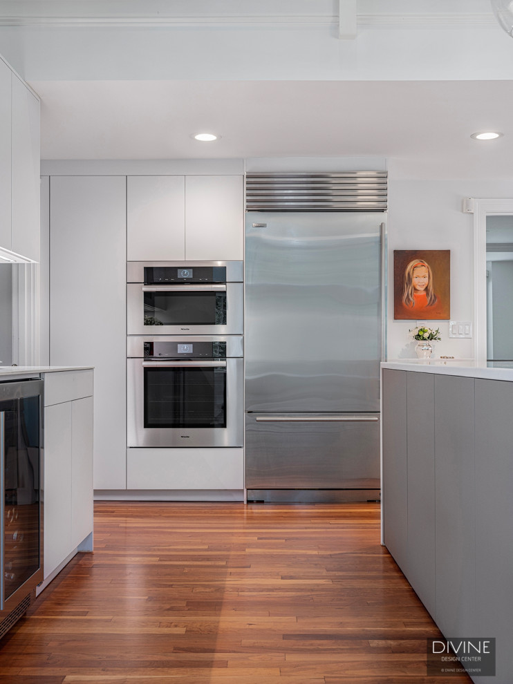 Inredning av ett modernt mellanstort vit vitt kök, med en undermonterad diskho, släta luckor, vita skåp, vitt stänkskydd, rostfria vitvaror, mellanmörkt trägolv, en halv köksö och brunt golv