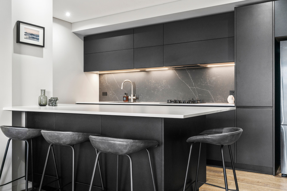 Mid-sized modern eat-in kitchen in Sydney with a drop-in sink, flat-panel cabinets, black cabinets, solid surface benchtops, black splashback, stone slab splashback, stainless steel appliances, medium hardwood floors, with island, brown floor and white benchtop.