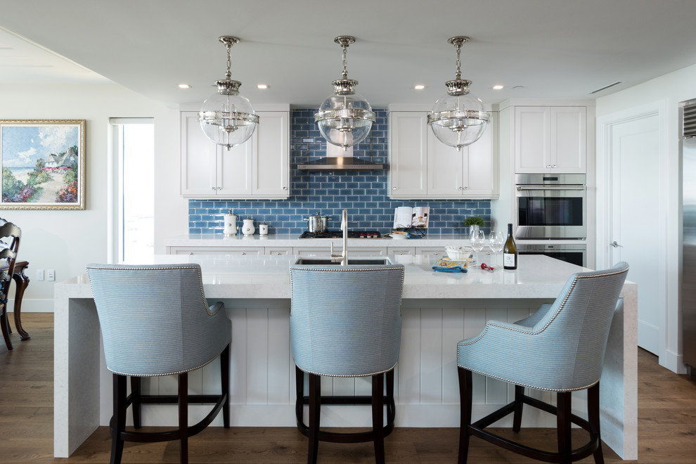 This is an example of a large beach style l-shaped eat-in kitchen in Miami with an undermount sink, shaker cabinets, white cabinets, blue splashback, subway tile splashback, stainless steel appliances, medium hardwood floors, with island, brown floor, white benchtop and granite benchtops.
