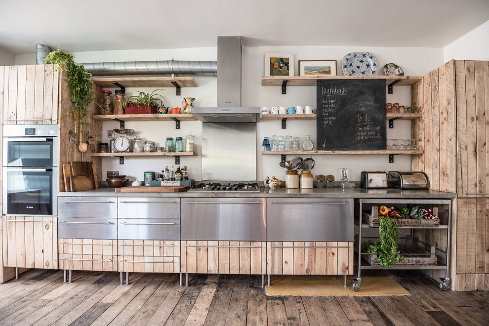 This is an example of an industrial single-wall kitchen in London with light wood cabinets, concrete benchtops, metallic splashback, stainless steel appliances, medium hardwood floors, no island and grey benchtop.